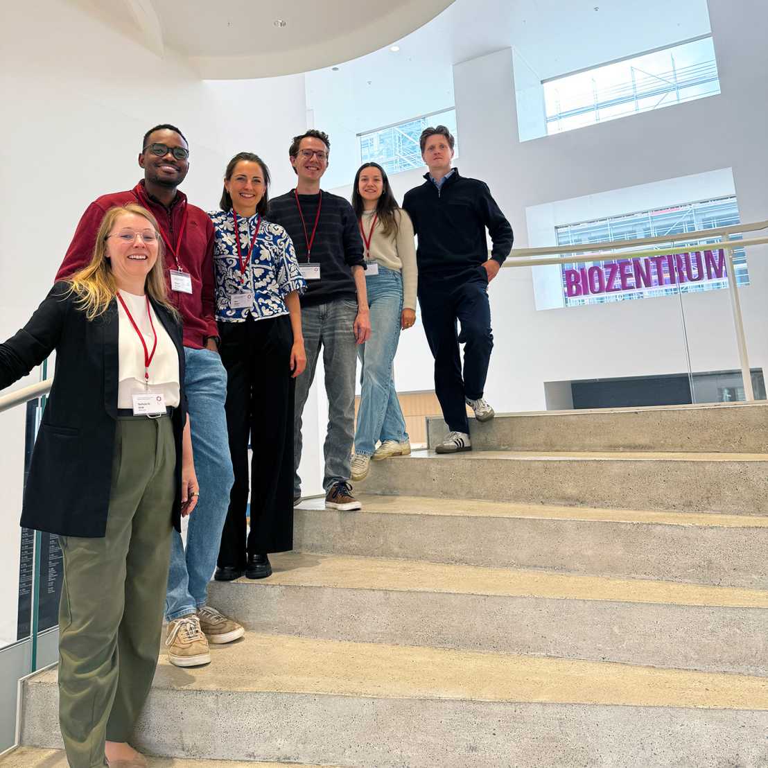 A group of six people standing on different steps of a stair with a sign in the backrgound reading Biozentrum